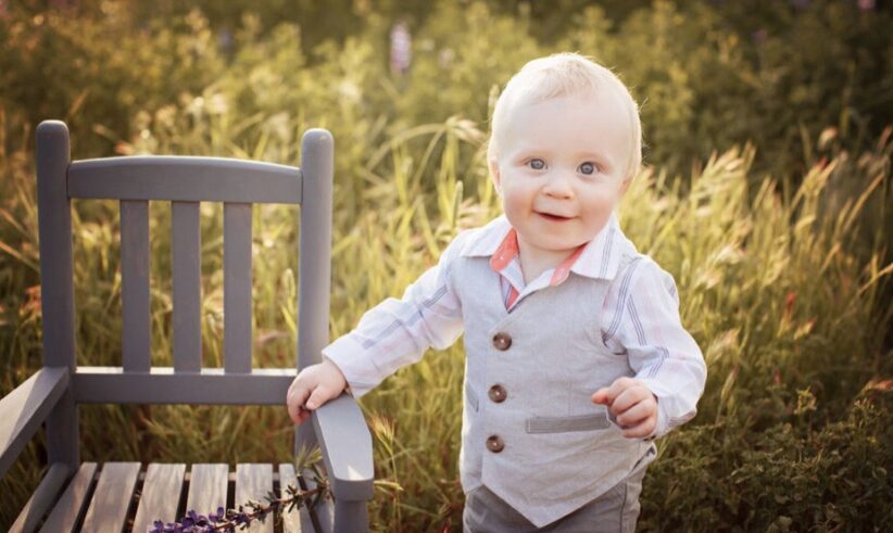 A smiling toddler with blond hair stands next to a gray wooden chair in a sunlit field, wearing a light gray vest, striped shirt, and pink collar. The background is filled with tall green grass and wildflowers.