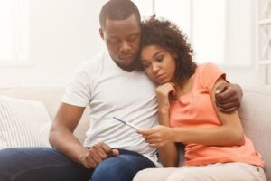 A couple sits close together on a couch, looking thoughtfully at a pregnancy test in the womans hand. The man has his arm around her shoulders, offering comfort as they both appear serious and contemplative.