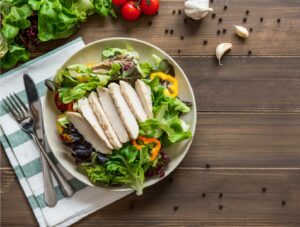A plate of salad topped with sliced grilled chicken breast, mixed greens, and colorful bell peppers sits on a wooden table with cutlery, a striped napkin, tomatoes, garlic cloves, and peppercorns nearby.