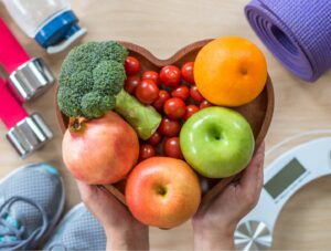 Hands hold a heart-shaped bowl filled with fresh fruits and vegetables, surrounded by fitness items including a yoga mat, dumbbells, sneakers, a water bottle, and a bathroom scale.