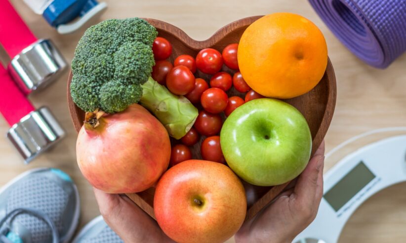 Hands hold a heart-shaped bowl filled with fresh fruits and vegetables, surrounded by fitness items including a yoga mat, dumbbells, sneakers, a water bottle, and a bathroom scale.