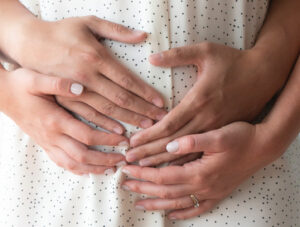 Close-up of four hands gently resting on a person’s belly, who is wearing a white shirt with black dots. Two hands are placed over two others, symbolizing care, support, or pregnancy.