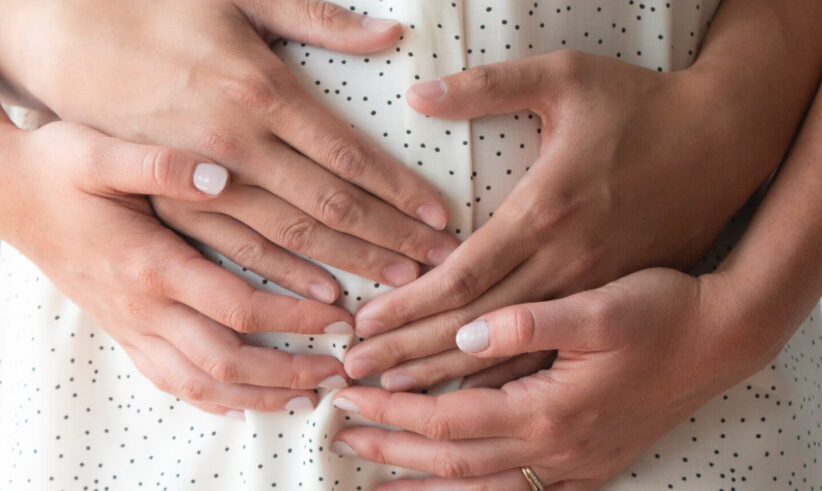 Close-up of four hands gently resting on a person’s belly, who is wearing a white shirt with black dots. Two hands are placed over two others, symbolizing care, support, or pregnancy.