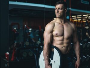 A muscular, shirtless man stands in a gym holding a weight plate in one hand, looking to the side. Gym equipment and a mirrored wall are visible in the background.