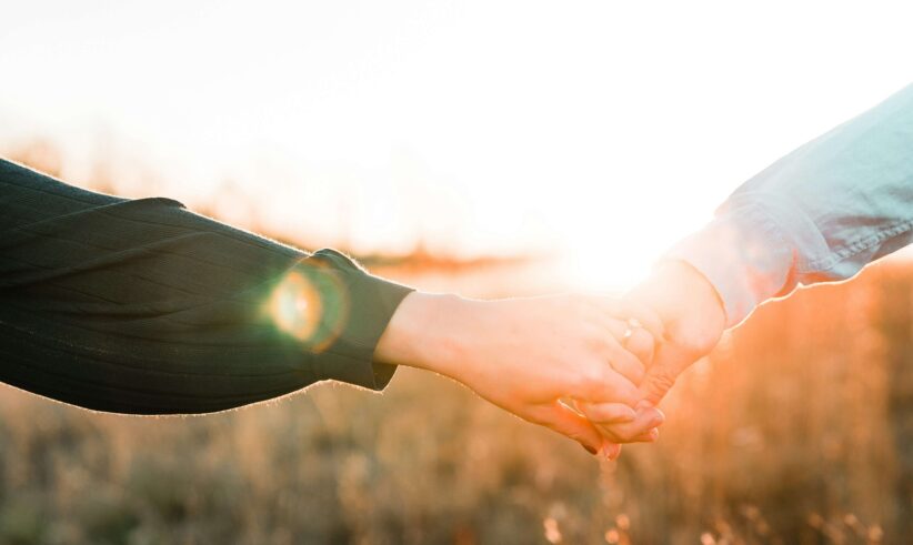Two people holding hands outdoors at sunset, with warm sunlight creating a soft, glowing effect in the background.