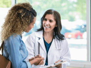 A smiling doctor in a white coat talks with a patient, holding a clipboard. They are standing indoors near large windows with sunlight coming in. The patient listens attentively and both appear engaged in conversation.