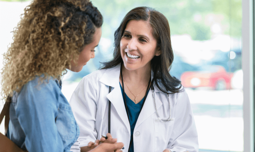 A smiling doctor in a white coat talks with a patient, holding a clipboard. They are standing indoors near large windows with sunlight coming in. The patient listens attentively and both appear engaged in conversation.