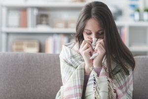 A young woman wrapped in a plaid blanket sits on a couch, holding a tissue to her nose as if she is sneezing or has a cold. The background shows a softly focused living room.