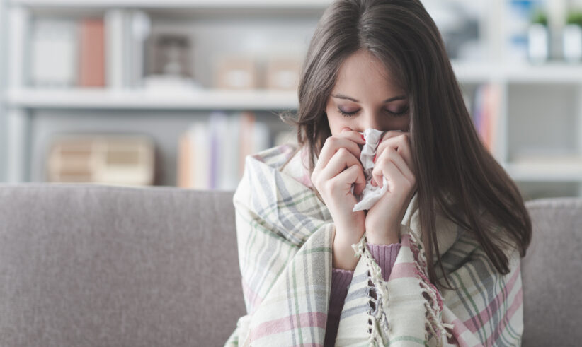 A young woman wrapped in a plaid blanket sits on a couch, holding a tissue to her nose as if she is sneezing or has a cold. The background shows a softly focused living room.