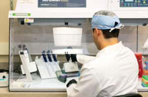 A laboratory technician wearing a lab coat, gloves, and a hair cover works under a fume hood, using a pipette to dispense liquid into a tray with several pipettes and lab equipment nearby.