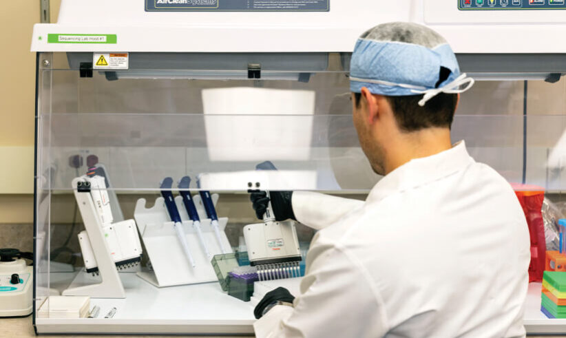 A laboratory technician wearing a lab coat, gloves, and a hair cover works under a fume hood, using a pipette to dispense liquid into a tray with several pipettes and lab equipment nearby.