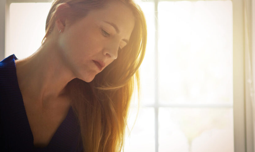 A woman with long red hair looks down thoughtfully, softly lit by sunlight coming through a window behind her. She wears a dark top, and the background is blurred and bright.