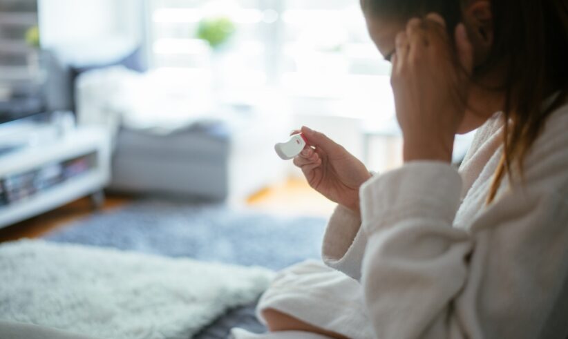 A person in a white robe sits indoors, holding a white electronic device and touching their forehead, with a blurred living room in the background.