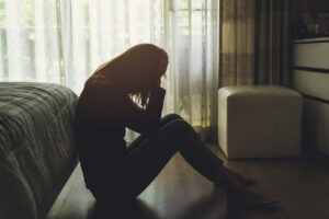 A person sits on the floor by a bed, silhouetted in natural light from a window, holding their head in their hands, appearing distressed or upset in a dimly lit room.
