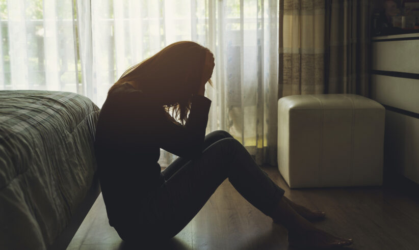 A person sits on the floor by a bed, silhouetted in natural light from a window, holding their head in their hands, appearing distressed or upset in a dimly lit room.