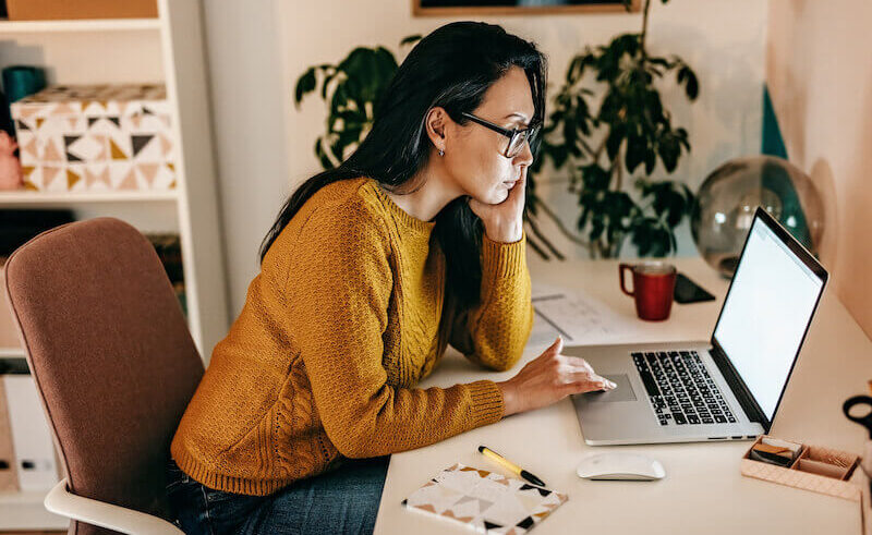 A woman wearing glasses and a yellow sweater sits at a desk, looking at a laptop screen. She rests her head on one hand, with a notebook, pen, and red mug nearby. The room is warmly lit with plants in the background.