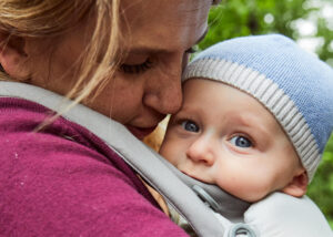 A close-up of an adult gently cuddling a baby in a carrier outdoors. The baby, wearing a blue knit hat, looks ahead with wide blue eyes while the adult’s face is lovingly pressed close to the babys head.
