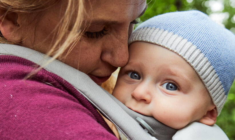 A close-up of an adult gently cuddling a baby in a carrier outdoors. The baby, wearing a blue knit hat, looks ahead with wide blue eyes while the adult’s face is lovingly pressed close to the babys head.