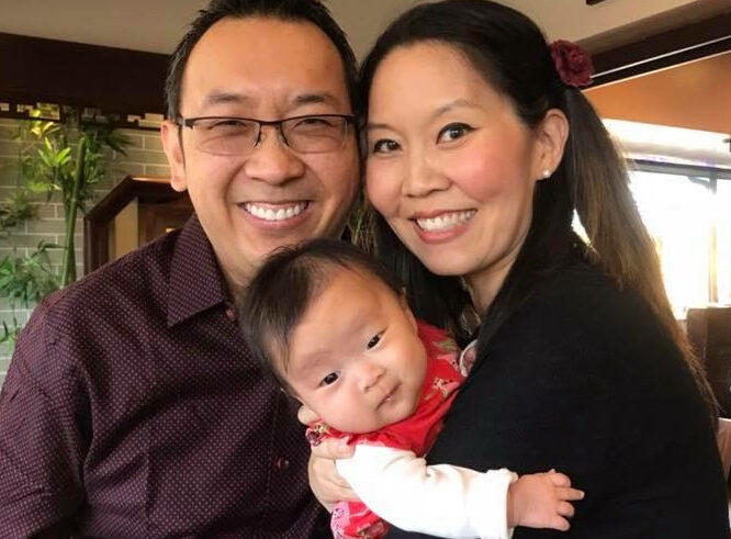 A smiling couple poses together indoors while holding a baby dressed in red. The man wears glasses and a maroon shirt; the woman has long dark hair, styled with a flower, and is wearing a black sweater.