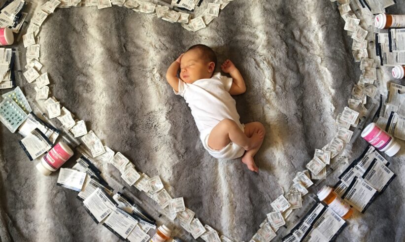 A newborn baby in a white onesie lies on a gray blanket, surrounded by a large heart shape made from medical supplies, including pill bottles, medication packets, and syringes.