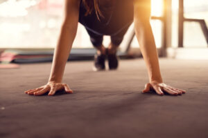 A person in athletic wear performing a push-up on a gym floor with their hands spread wide, focused on exercise. Sunlight streams in from windows in the background.