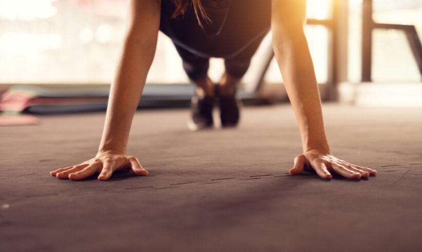 A person in athletic wear performing a push-up on a gym floor with their hands spread wide, focused on exercise. Sunlight streams in from windows in the background.