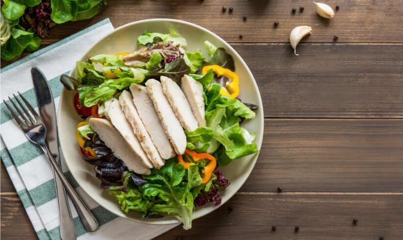 A plate of fresh salad with sliced grilled chicken breast, mixed greens, yellow bell pepper, and cherry tomatoes on a wooden table, with utensils and a napkin beside it. Garlic, tomatoes, and peppercorns are scattered nearby.