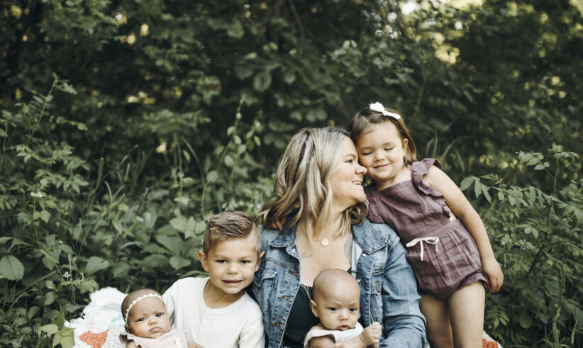 A woman sits on a quilt outdoors with four young children—two babies in her lap, a toddler girl standing and hugging her, and a young boy sitting close—surrounded by lush greenery.