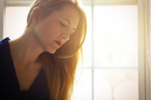 A woman with long light brown hair looks down thoughtfully, illuminated by soft sunlight streaming through a window in the background. She wears a dark top and appears to be indoors.