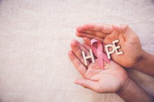 Two cupped hands hold wooden letters spelling HOPE, with a pink ribbon replacing the O, symbolizing breast cancer awareness and hope. The background is a neutral, textured fabric.