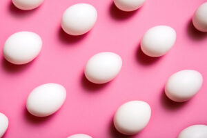 White eggs are arranged in a diagonal grid pattern on a bright pink background. The eggs are evenly spaced and their shadows create a sense of depth.