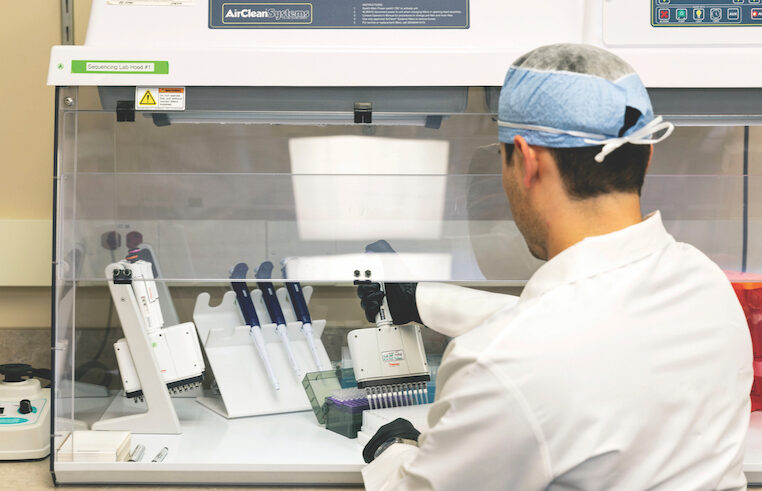 A lab technician in protective clothing works at a biosafety cabinet, using pipettes and handling samples with lab equipment in a sterile environment.