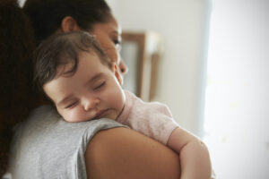A baby is sleeping peacefully on an adult’s shoulder. The adult is facing away from the camera, gently holding the baby. Soft natural light fills the calm, cozy room.
