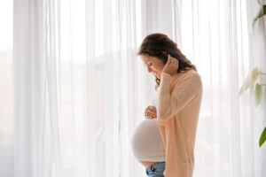 A pregnant woman standing by a window with sheer white curtains, gently touching her belly and looking down, wearing a light-colored top and cardigan in a softly lit room.