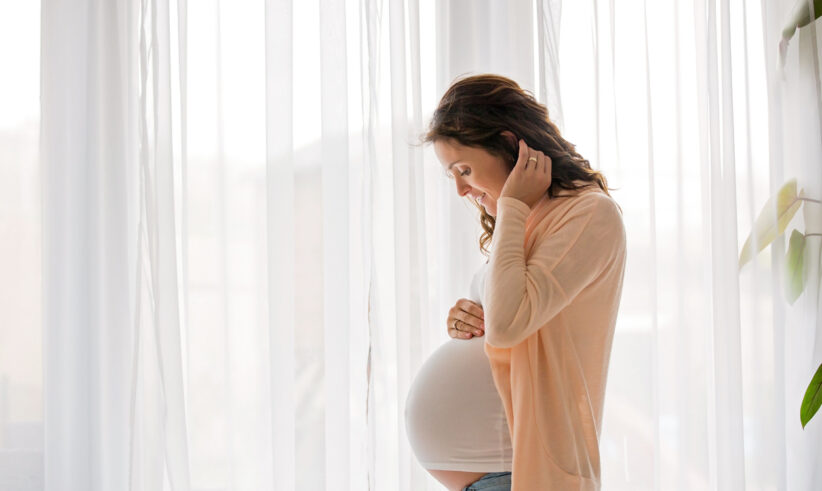 A pregnant woman standing by a window with sheer white curtains, gently touching her belly and looking down, wearing a light-colored top and cardigan in a softly lit room.