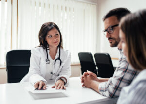 A female doctor in a white coat with a stethoscope talks to a man and a woman sitting across from her at a desk in a medical office with large windows and blinds.