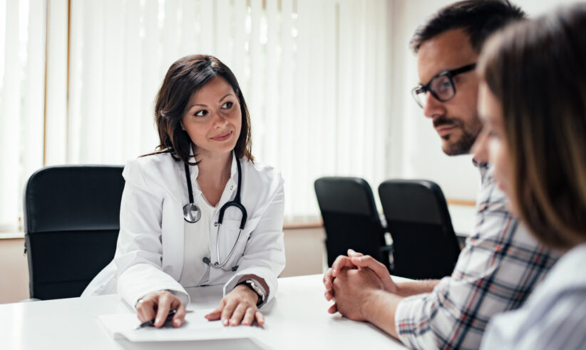 A female doctor in a white coat with a stethoscope talks to a man and a woman sitting across from her at a desk in a medical office with large windows and blinds.