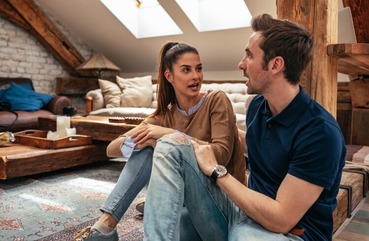 A woman and a man sit on the floor of a cozy living room, engaged in conversation. The woman has her hair in a ponytail and gestures with her hand, while the man listens attentively. Warm light fills the room.