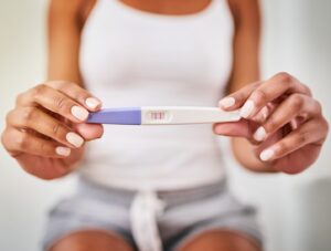A person wearing a white tank top and shorts holds a pregnancy test with two red lines, indicating a positive result. The focus is on the test and their hands.