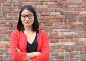A woman with straight black hair and glasses stands in front of a brick wall, wearing a red jacket over a black top, with her arms crossed and a slight smile.