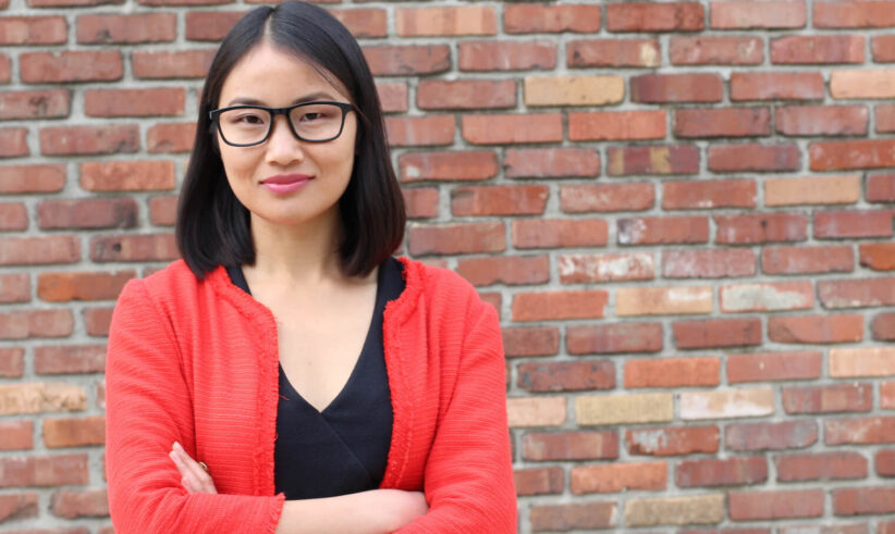 A woman with straight black hair and glasses stands in front of a brick wall, wearing a red jacket over a black top, with her arms crossed and a slight smile.