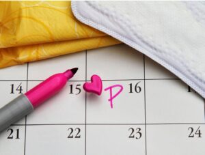 A pink marker, heart-shaped pin, and the letter P mark the 15th on a calendar, with a sanitary pad and yellow wrapper nearby, symbolizing a menstrual period reminder.