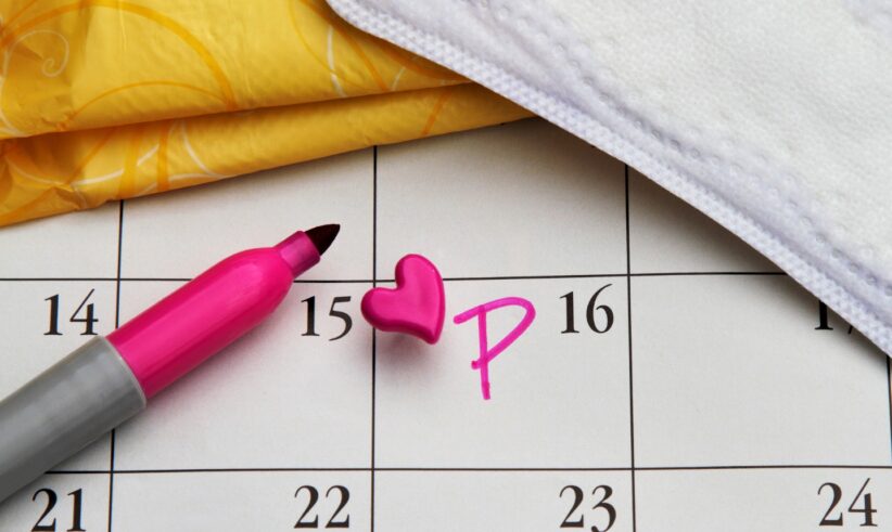 A pink marker, heart-shaped pin, and the letter P mark the 15th on a calendar, with a sanitary pad and yellow wrapper nearby, symbolizing a menstrual period reminder.