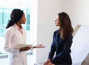A doctor in a white coat speaks to a woman sitting on an exam table in a medical office, holding a file while the patient listens attentively.