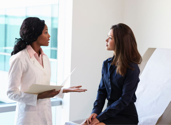 A doctor in a white coat speaks to a woman sitting on an exam table in a medical office, holding a file while the patient listens attentively.