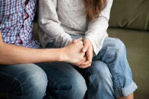 Two people sitting closely on a couch, holding hands in a comforting gesture. One wears a plaid shirt and jeans, the other a gray long-sleeve top and jeans. Their faces are not visible.