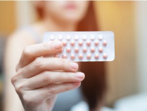 A person holds a blister pack of oral contraceptive pills, with pink and white tablets visible, against a blurred indoor background.