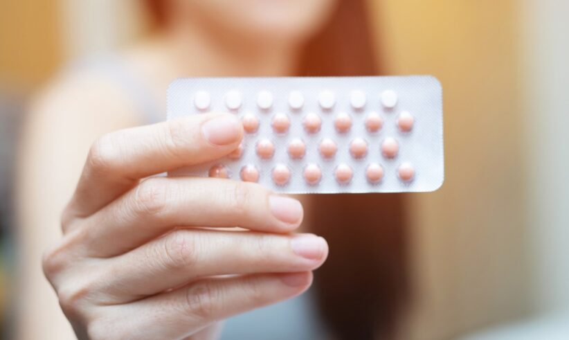 A person holds a blister pack of oral contraceptive pills, with pink and white tablets visible, against a blurred indoor background.