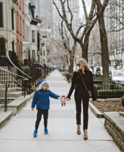 A woman in black attire and sunglasses walks on a city sidewalk, holding hands with a young boy in a blue coat and hood. The boy is holding a small stuffed toy. Leafless trees line the street on an overcast day.