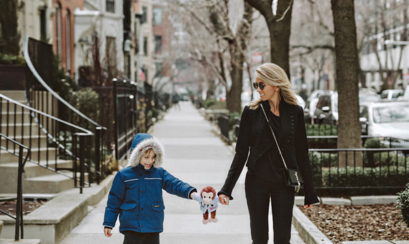 A woman in black attire and sunglasses walks on a city sidewalk, holding hands with a young boy in a blue coat and hood. The boy is holding a small stuffed toy. Leafless trees line the street on an overcast day.